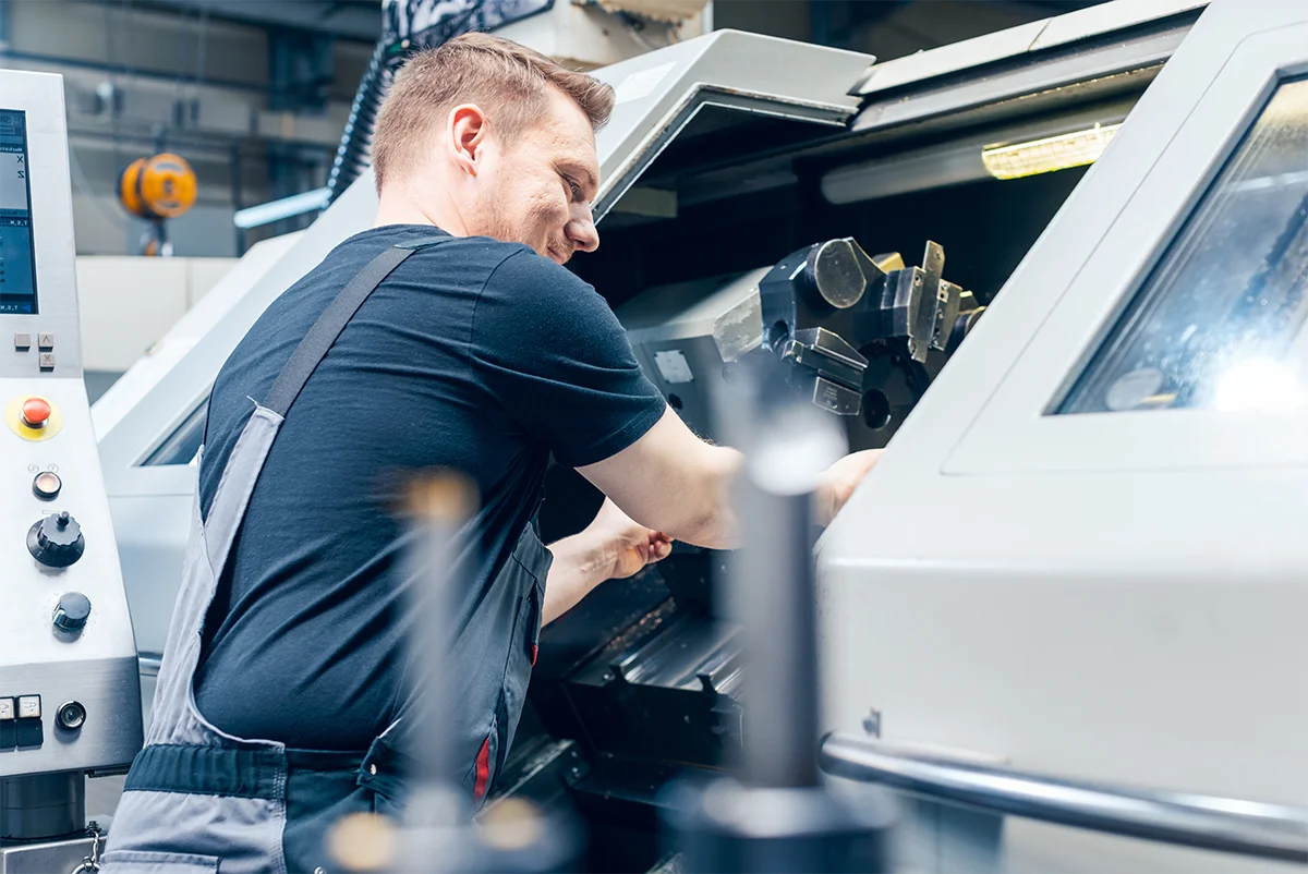 Eine Person im Overall arbeitet in einer Werkstatt an einer CNC-Maschine. Sie ist konzentriert auf die Justierung von Teilen im Inneren der Maschine konzentriert. Werkzeuge und Bedienfelder sind in der Nähe sichtbar. Die Umgebung wirkt industriell, das Licht spiegelt sich in den Metalloberflächen.