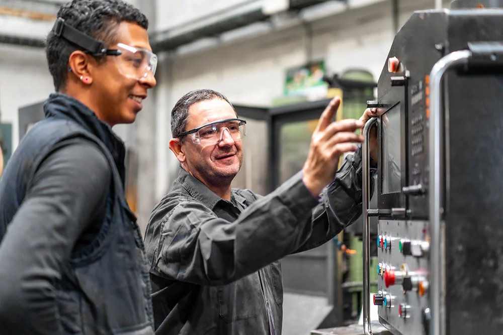 Zwei Personen mit Schutzbrillen arbeiten an einem Bedienfeld in einer Industrieanlage. Eine Person zeigt auf den Bildschirm der Maschine, während die andere zusieht. Beide tragen dunkle Arbeitskleidung.