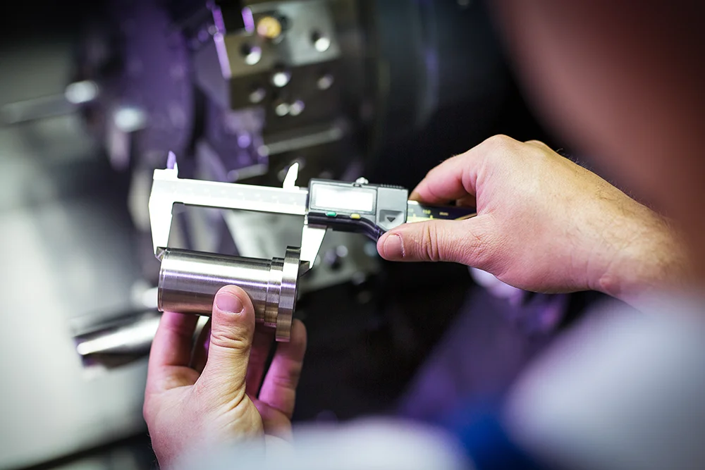 Eine Person misst in einer Werkstatt mit einem digitalen Messschieber den Durchmesser eines zylindrischen Metallteils. Der Hintergrund ist unscharf und fokussiert den Präzisionsmessvorgang.