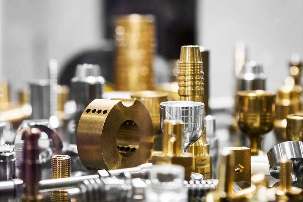 Nahaufnahme verschiedener präzisionsgefertigter Metallkomponenten, hauptsächlich in Gold- und Silberfarben. Die Teile haben unterschiedliche Formen und Texturen, darunter zylindrische und zahnradartige Designs, und sind in einer gruppierten Darstellung angeordnet.